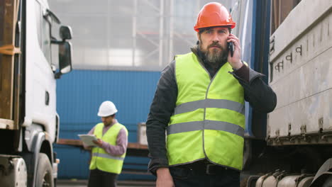 Arbeiter-Mit-Weste-Und-Schutzhelm,-Der-Eine-LKW-Flotte-In-Einem-Logistikpark-Organisiert,-Während-Er-Telefoniert-1