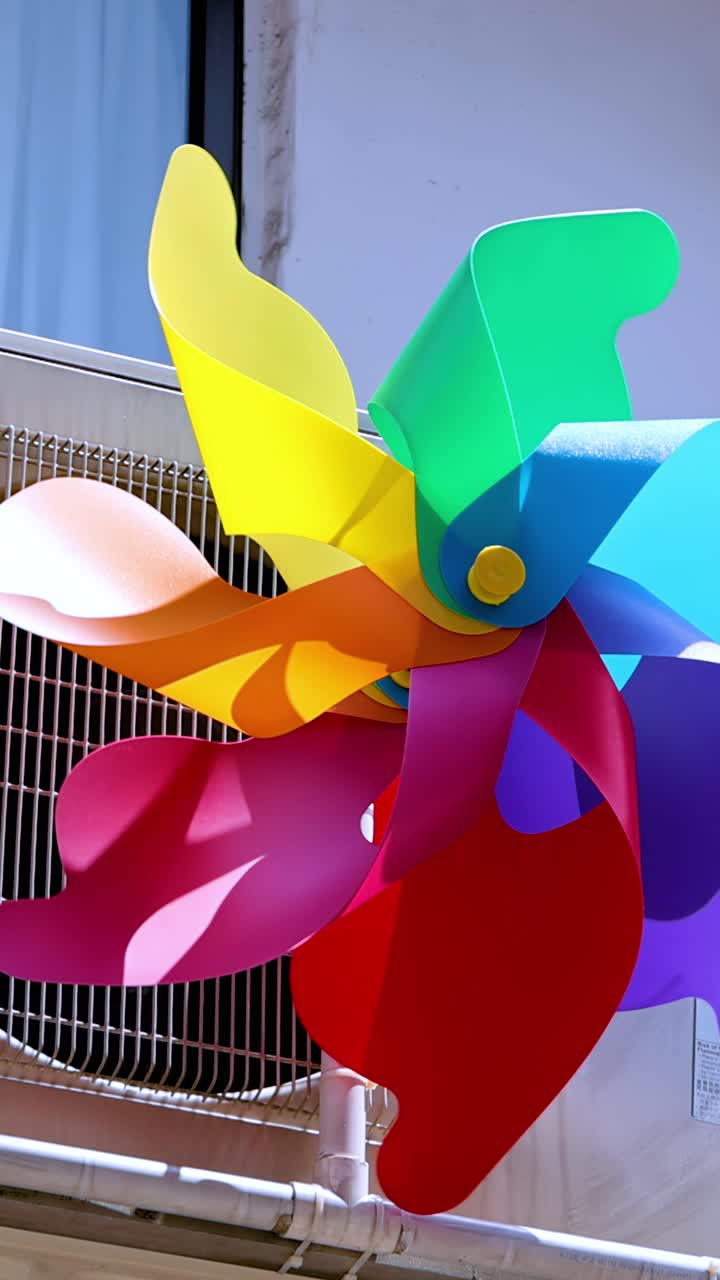 Un vibrante molinillo de viento de plástico gira rápidamente en un balcón en Singapur, iluminado por una fuerte luz diurna. La cámara permanece estable, capturando el movimiento dinámico y los colores vivos.