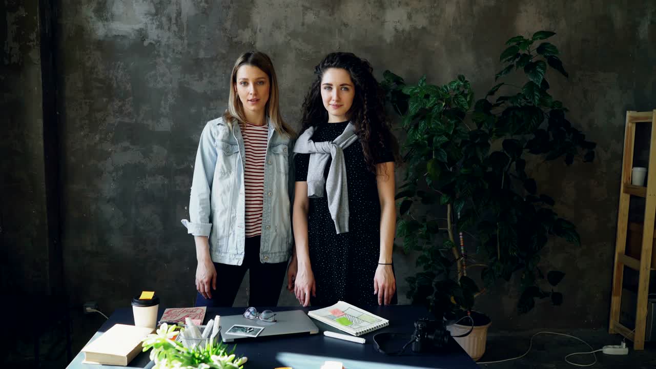 Two young female business colleagues are standing together in loft style office, posing and looking at camera. Girls are happy, smiling and laughing