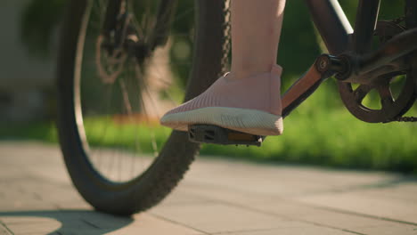 close-up of leg in pink sneakers tapping bicycle pedal while other foot remains grounded, preparing for ride, casting shadow on ground with blurred greenery in background