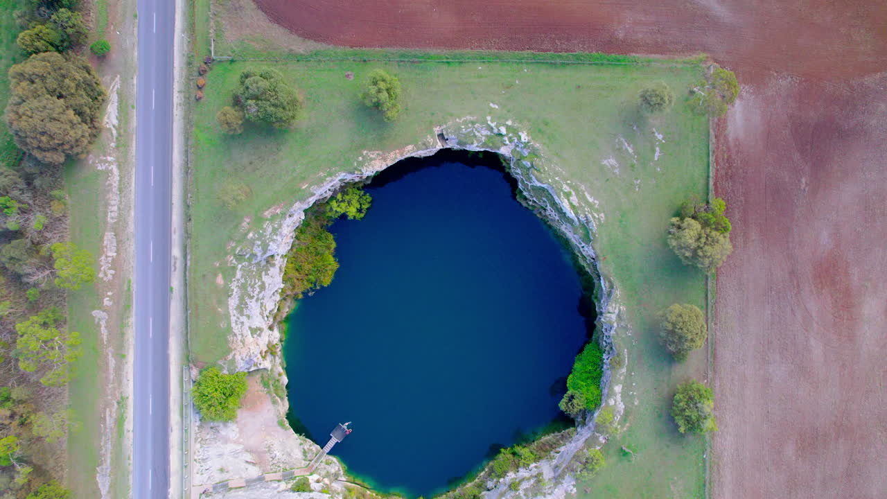Top-down drone shot of Little Blue Lake in South Australia, revealing its striking blue water surrounded by green and brown fields in a remote rural landscape