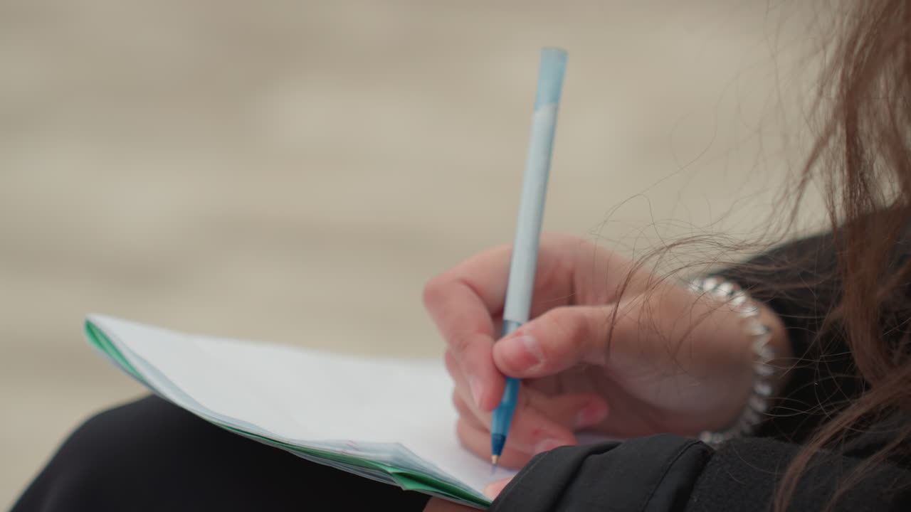 student journaling on pad in urban setting, closeup of hand holding pale blue pen, spiral pad on lap, blurred pavement background, casual bracelet, slow creative brainstorming, relaxed study break