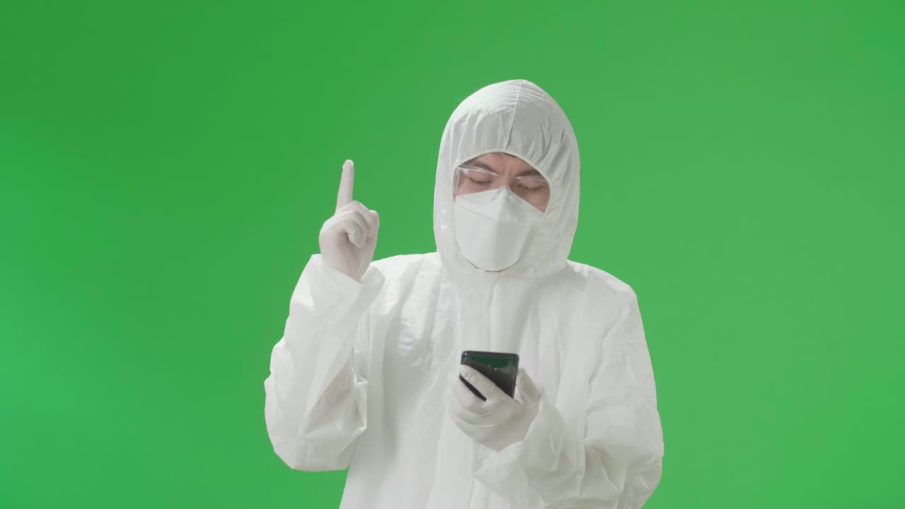 Asian Man Wearing Personal Protective Equipment Uniform Ppe, Medical Face Mask And Use Mobile Phone Then Thinking About Something, Looking Around Then Raising His Index Finger In The Green Screen Studio