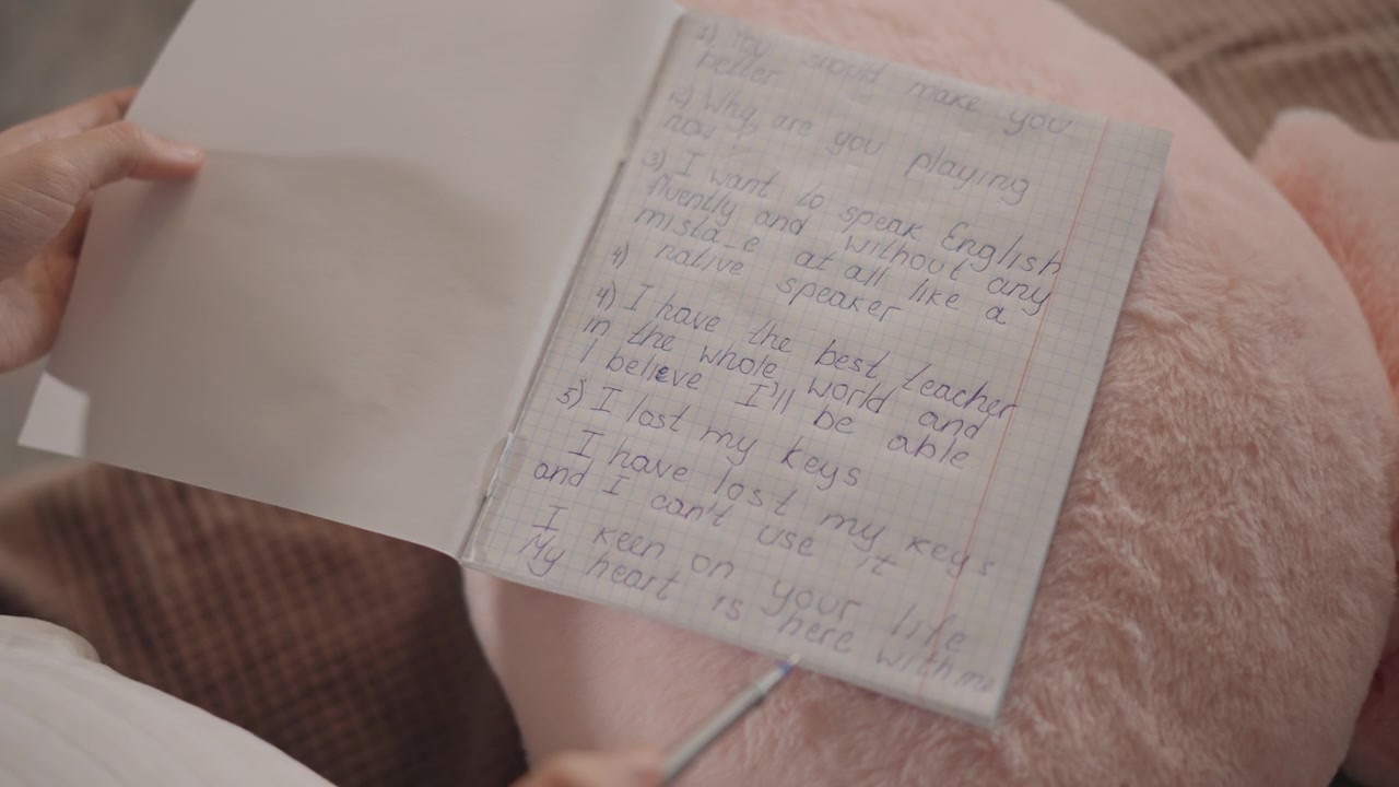 Top-down view of person holding open notebook with handwritten notes, left hand flipping page while right hand keeps pen poised, capturing steady writing preparation and quiet study mood indoors
