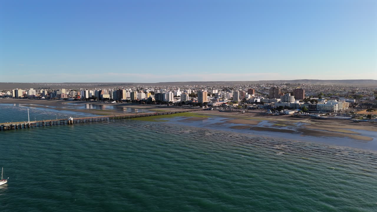 Coastal aerial of Puerto Madryn’s pier, vibrant city and ocean view under a sunny sky, dynamic tourism hub, establishing