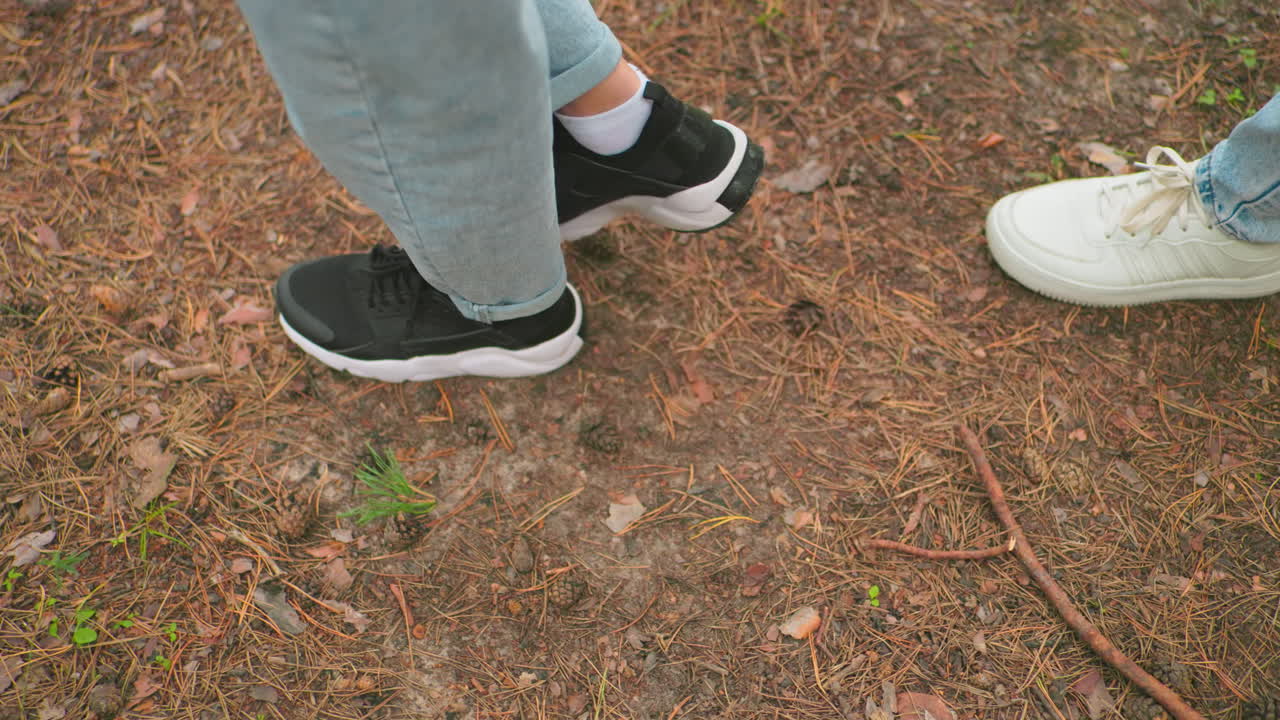 Close-up of two individuals walking on a forest trail wearing blue jeans, with one in black sneakers and the other in white sneakers, surrounded by pine needles, leaves, and small twigs