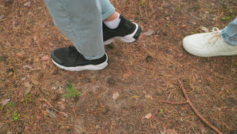 close-up of two individuals walking on a forest trail wearing blue jeans, with one in black sneakers and the other in white sneakers, surrounded by pine needles, leaves, and small twigs