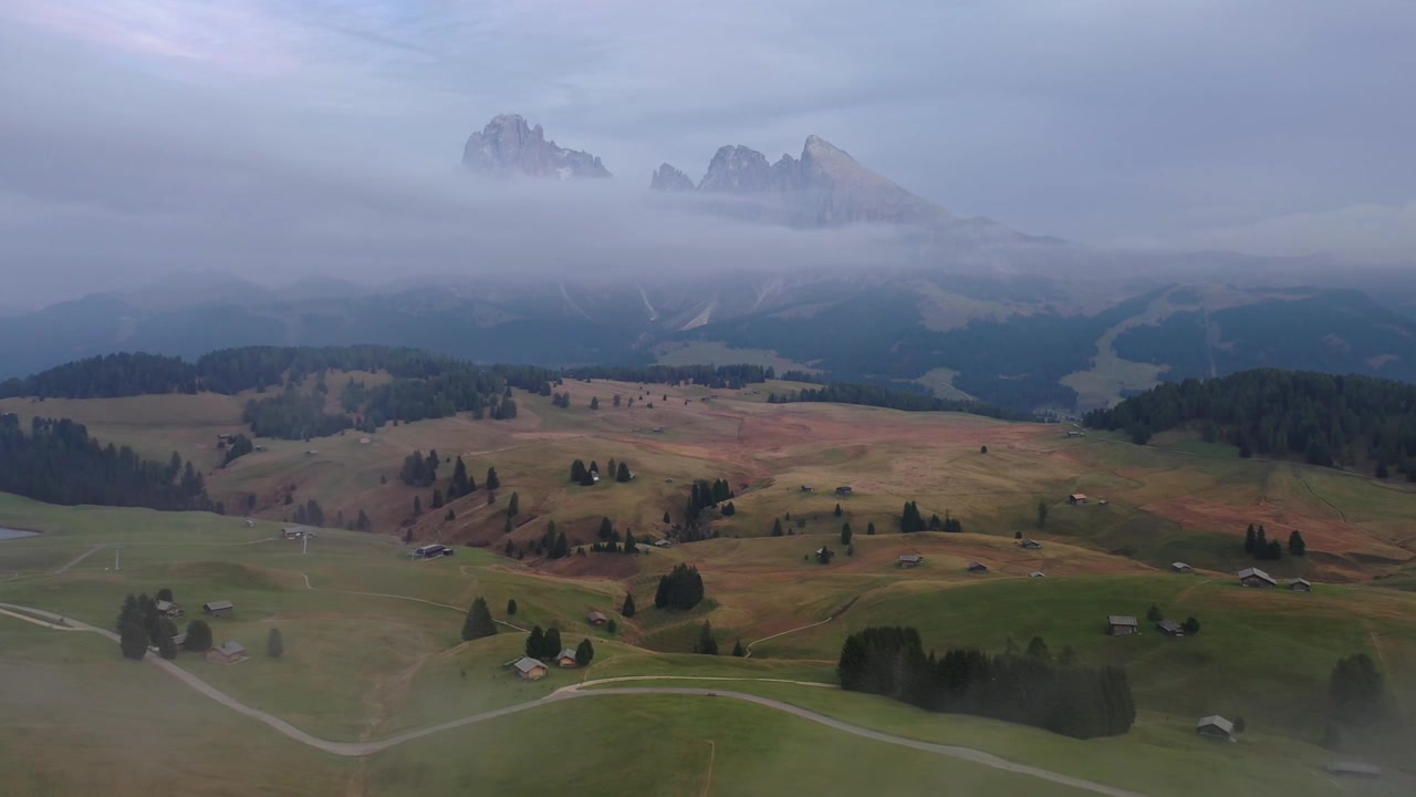 Imponerende droneoptagelse fra høj vinkel af tykke lavtliggende skyer og tåge, der ruller gennem Alpe di Siusi-dalen i Italien. Viser en grøn fyrreskovsryg med de majestætiske Sassolungo-toppe, der rejser sig over.