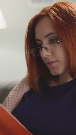 focused woman with ginger hair enjoys reading captivating detective story in evening. housewife holds red fiction book flipping pages at home closeup