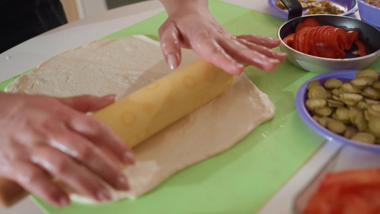 Primo piano dall'alto di mani che premono e fanno rotolare un mattarello di legno decorato su impasto appiattito su un tappetino di silicone verde brillante, mostrando un movimento avanti e indietro con la pressione del palmo che modella.