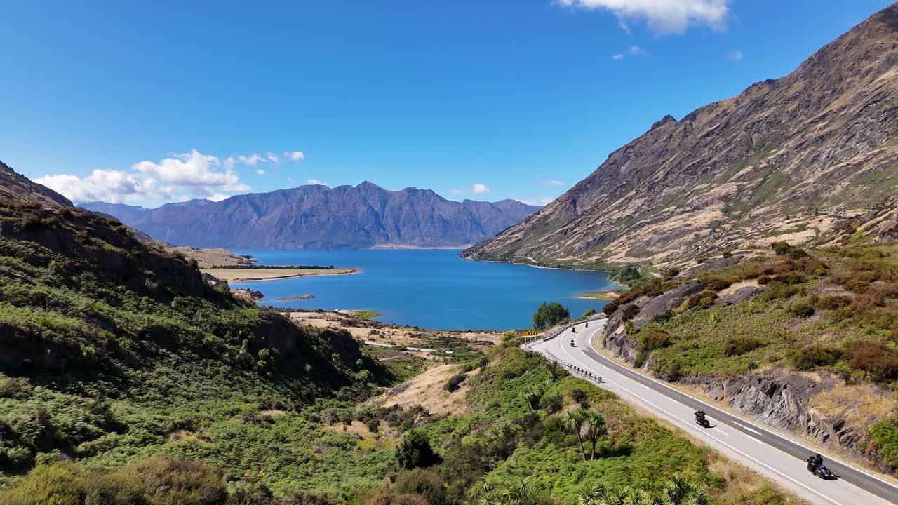 4k Drone video of a group of maori bikers driving along the road at Lake Hawea in Wanaka, New Zealand. The roads wind around hills and mountains next to the blue, vast and expansive lake