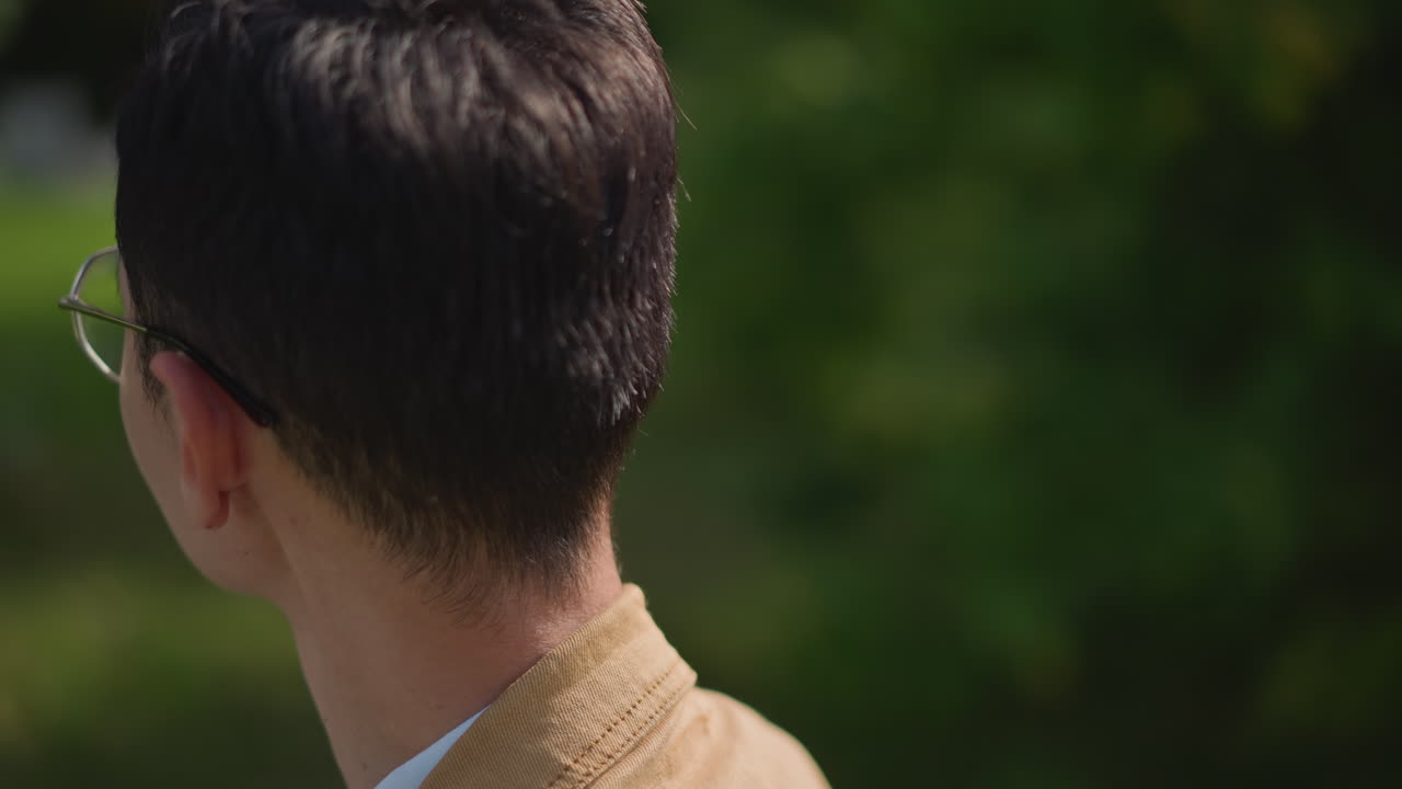 asian student on park pathway looking sideways, casual tan jacket and glasses catching light, brief walking pause with backview and profile frames, cinematic bokeh, reflective mood, campus street vibe