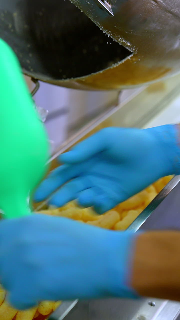Factory worker takes corn sticks out of a metal tank. Crispy puff corns are put on the conveyor belt for the next stage of production. Vertical video