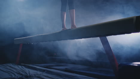 the girl is a professional athlete performs gymnastic acrobatic trick on a beam in backlight and slow motion in sports gymnastic clothing. smoke and blue. jump and spin on the balance beam