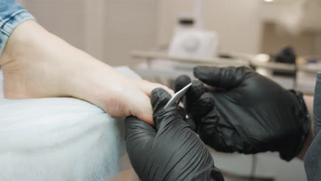 technician wearing black gloves carefully filing toenails of client lying on treatment chair, with tools and equipment visible in blurred background of clean, professional salon environment