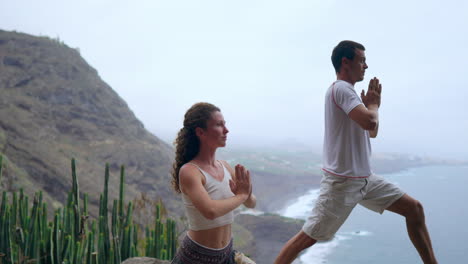 un hombre y una mujer de pie en el borde de un acantilado con vistas al océano levantan las manos y inhalan el aire del mar durante el yoga