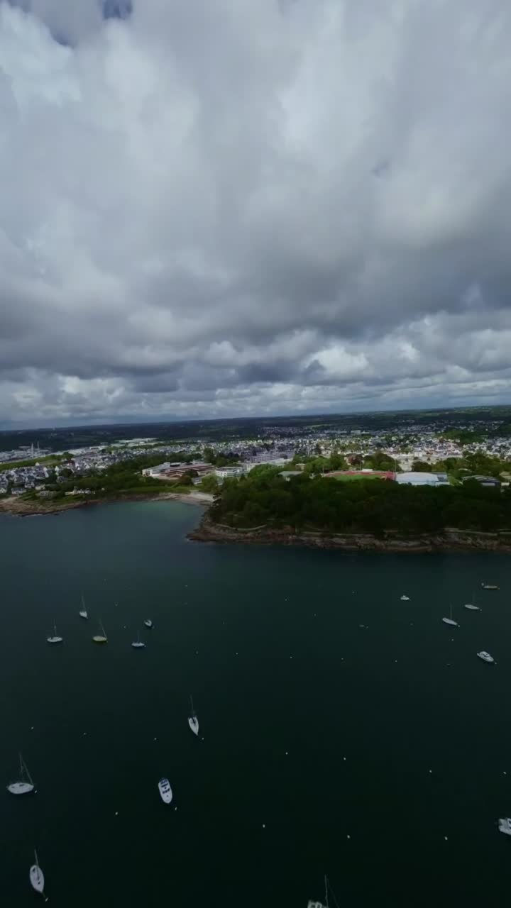 Aerial drone view France Atlantic peninsula charm, Brittany coastal town residential streets, sports field and boats in calm harbor under cloudy sky