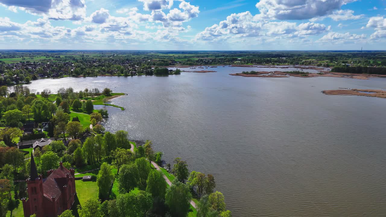 A lakeside village with a red-roofed church nestles among green trees, overlooking a wide calm lake beneath dramatic spring skies and billowing clouds.