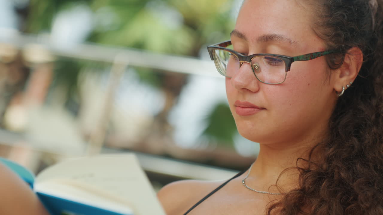Primer plano de mujer leyendo un libro al aire libre, retrato de estudiante reflexiva con gafas y cabello rizado, luz solar suave en la piel, fondo bokeh de hojas, sonrisa suave al pasar las páginas, ambiente de estudio relajado