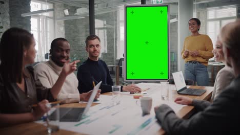 project manager makes a presentation on a green screen mock up display for a young diverse creative team in meeting room in an agency. colleagues sit behind conference table and discuss business.