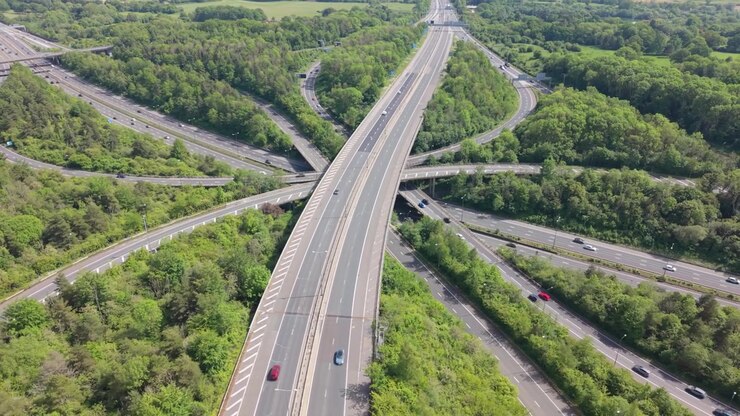 Aerial view of a major highway interchange surrounded by green forest. Cars and trucks drive on multiple elevated roads and ramps. Bright sunny day with flowing traffic. Busy transportation hub.