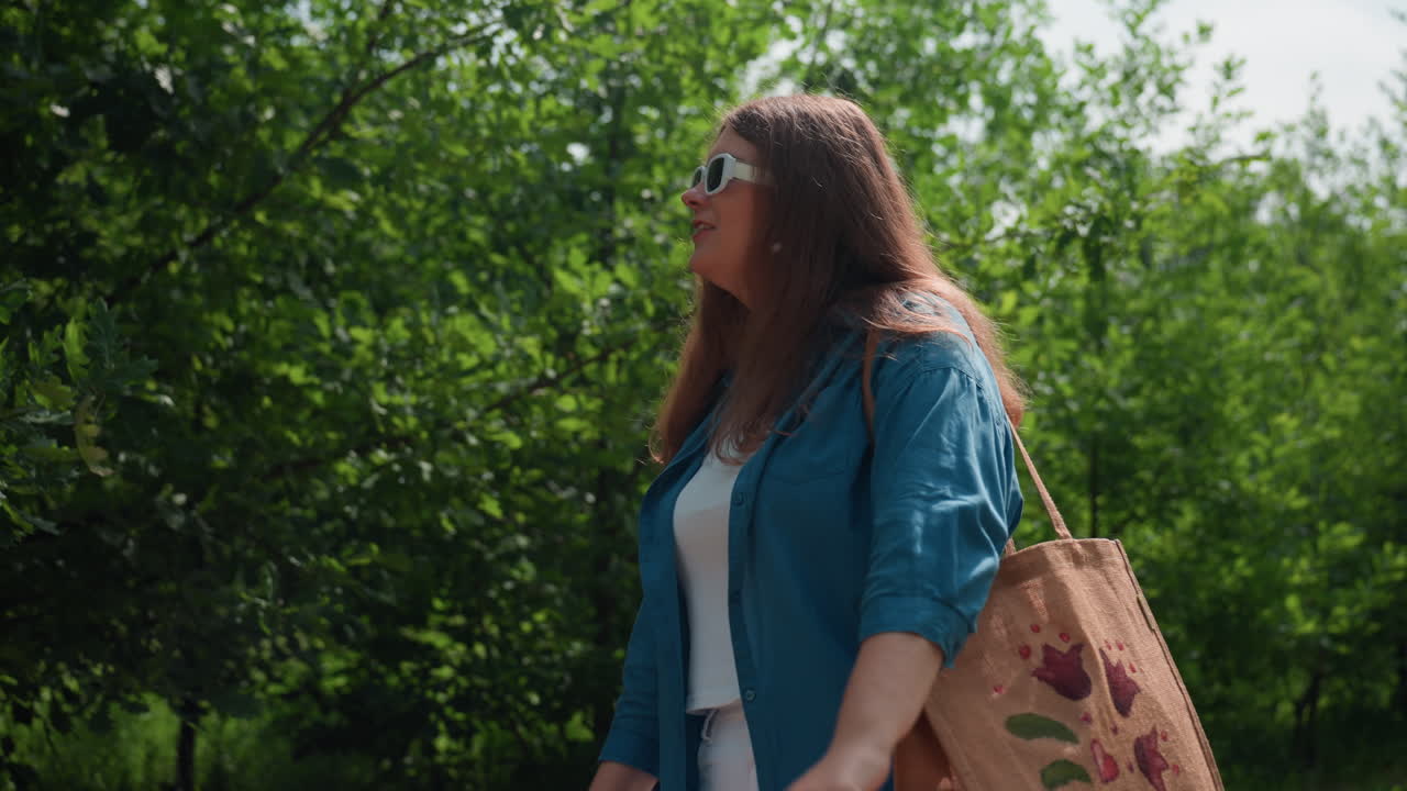 Una mujer baila alegremente en un sendero soleado de un parque con un bolso bordado al hombro, lleva una camisa azul y un conjunto blanco, rodeada de árboles verdes, disfrutando del ambiente veraniego.