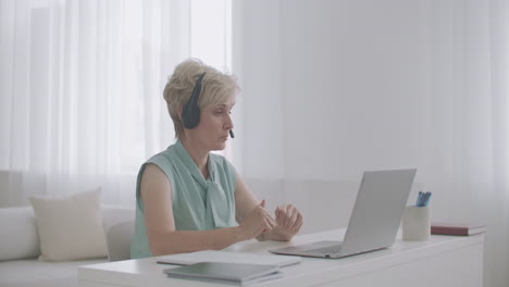 female online consultant is talking with client by headphones with microphone looking at screen of laptop working from home distant work