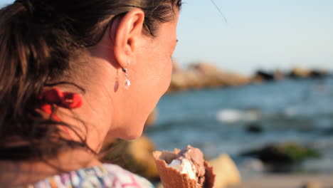 toma en cámara lenta de una hermosa mujer con un aspecto natural lamiendo un delicioso helado en la playa mientras las olas rompen y el sol ilumina su rostro