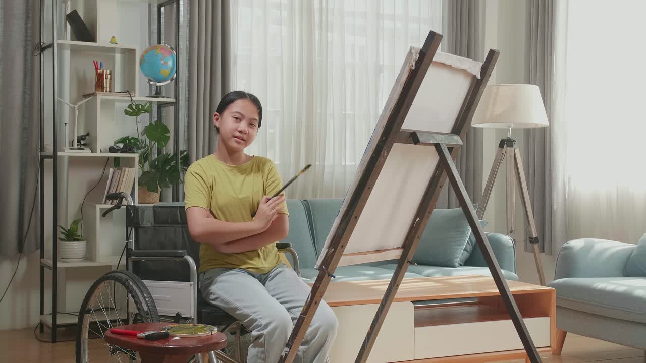 Asian Artist Girl In Wheelchair Holding Paintbrush And Cross His Arms And Smile While Painting On The Canvas