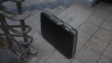 unattended black briefcase left on a concrete stairwell. suspicious placement, secret drop, espionage exchange, or potential threat. evokes tension, mystery, and cinematic intrigue. money bag