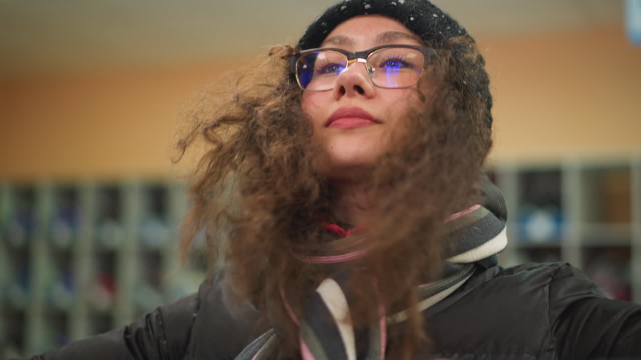 Damsel with long curly hair wearing black winter jacket and striped scarf tilts head slightly down,as she wears her head warmer soft indoor lighting highlighting casual natural moment