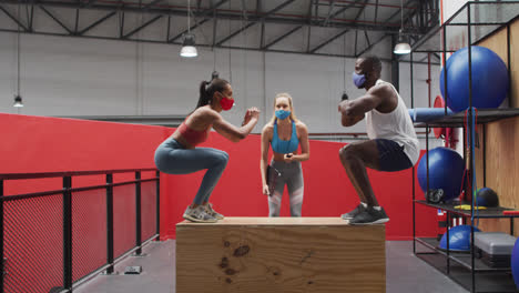 diverse man and woman jumping on box wearing face masks at gym, caucasian woman timing