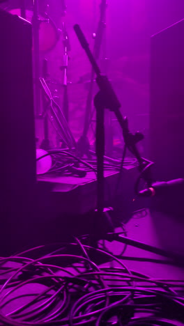a dimly lit stage with purple spotlights, microphones, and a tangle of cables. the image captures the backstage atmosphere of a concert or live event.