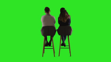 rear view studio shot of man and woman sitting on stools facing away from camera against green screen