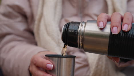close up of woman hands with french manicure pouring hot tea from stainless steel thermos into metal cup while relaxing on park bench amid vibrant autumn foliage wearing warm jacket and chunky scarf