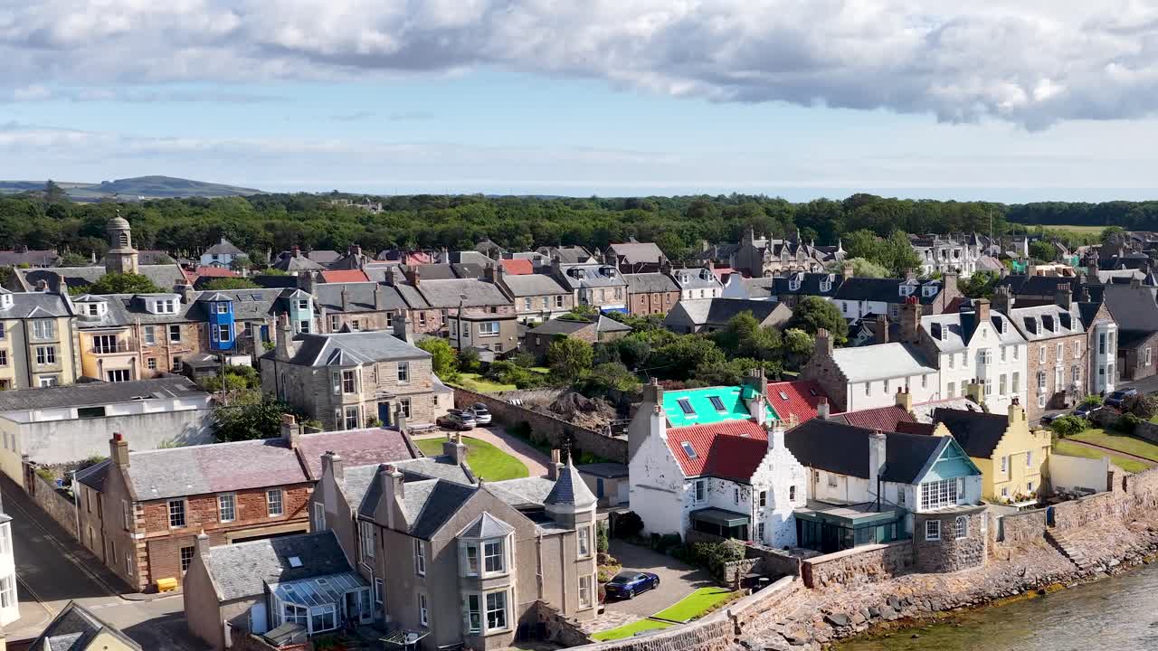 Drone camera glides above seaside rooftops, revealing historic architecture and lush landscape in daylight