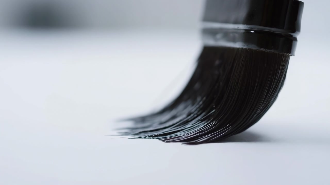 Primo piano ravvicinato a bassa altezza dalla superficie della punta di un pennello che preme su una superficie piatta e chiara e lascia una fresca pennellata nera curva; vernice viscosa bagnata ricopre le singole setole con una fine separazione e una leggera lucentezza.