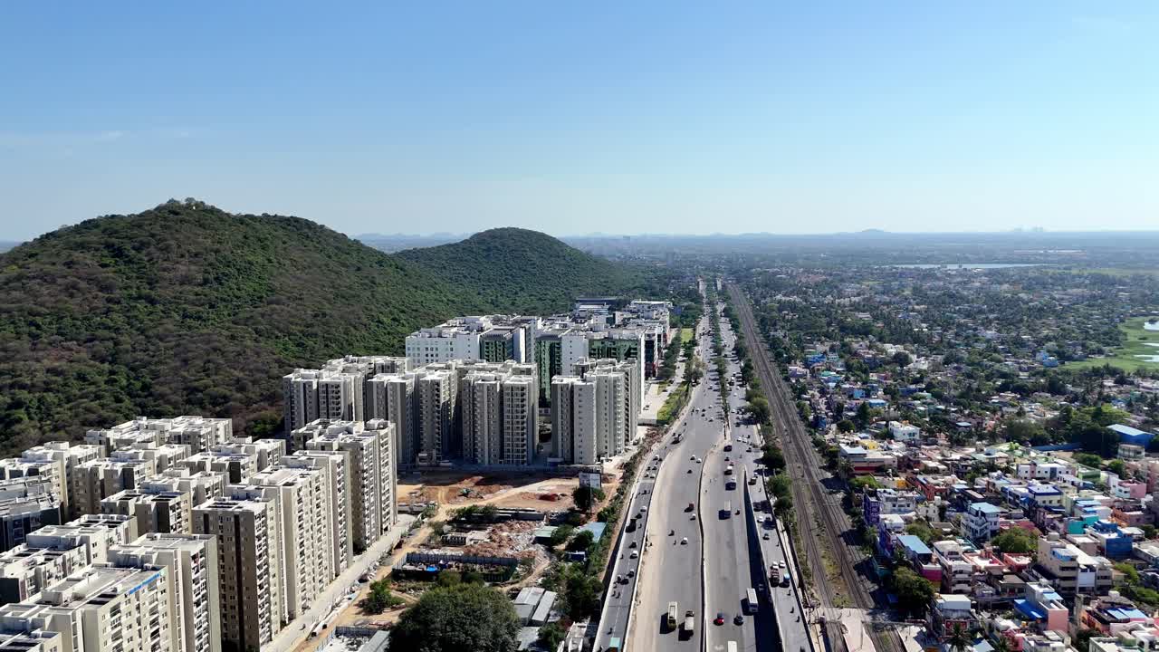 Dynamic aerial view of Chennai's urban growth. Features new high-rise residential buildings, a multi-lane highway, and parallel railway tracks. Green hills provide a natural backdrop contrasting.