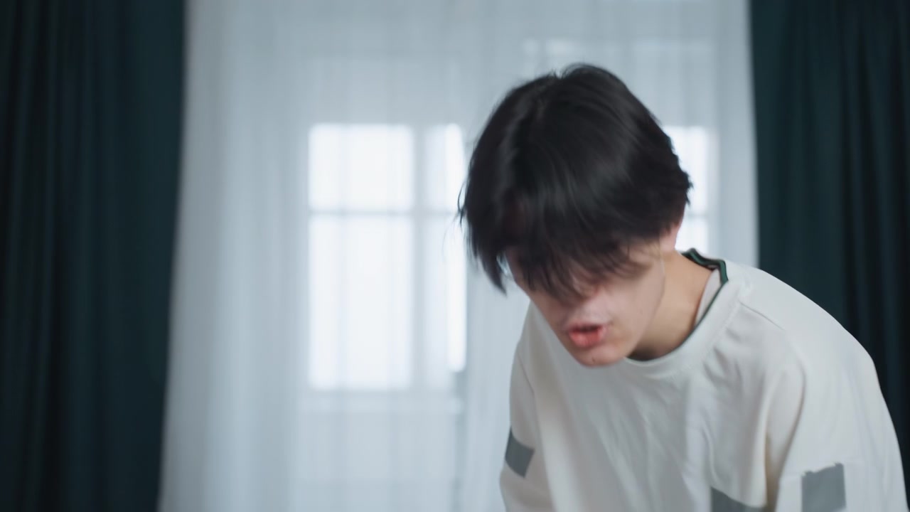 집에서 침실을 정리하는 젊은 아시아 남성. 얇은 커튼 옆 서랍장 위로 몸을 숙이고 있습니다. 부드러운 아침 햇살 아래 집중된 표정으로 천과 접힌 리넨을 부드럽게 닦는 동작을 합니다. 차분한 미니멀리스트 인테리어입니다.