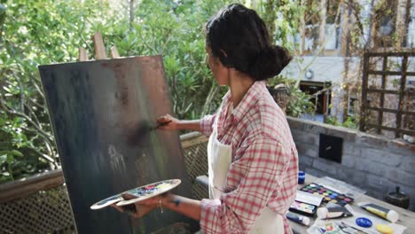 biracial female artist in apron painting on canvas in sunny garden, slow motion