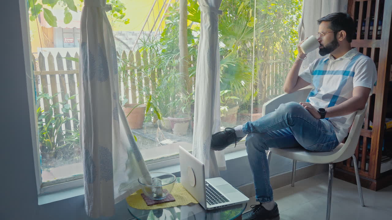 Full shot of a man drinking coffee near the window side and checking time in his smartwatch.