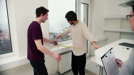 Male-couple-viewing-a-property-together-with-agent,-measuring-distance-at-kitchen