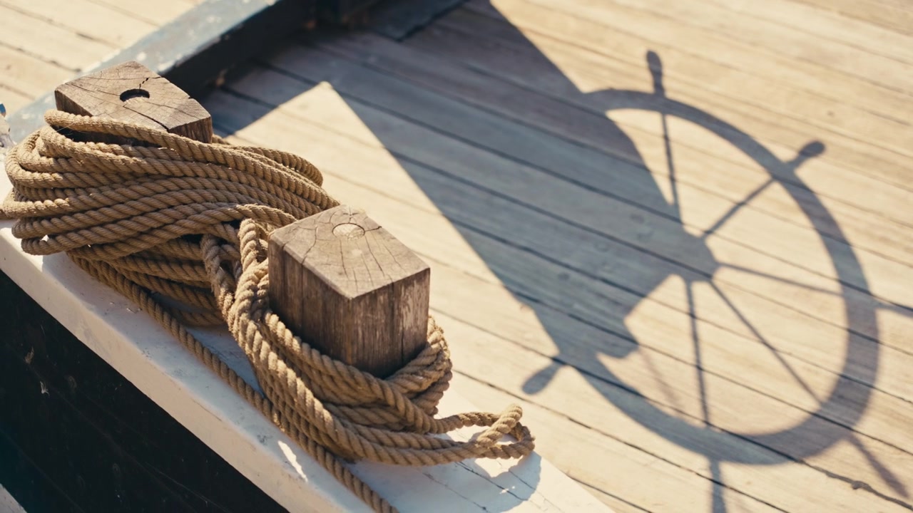 Smooth rack focus transitions from a steering wheel's silhouette cast on a weathered wooden ship deck to coiled nautical ropes wrapped around cleats in warm sunlight.