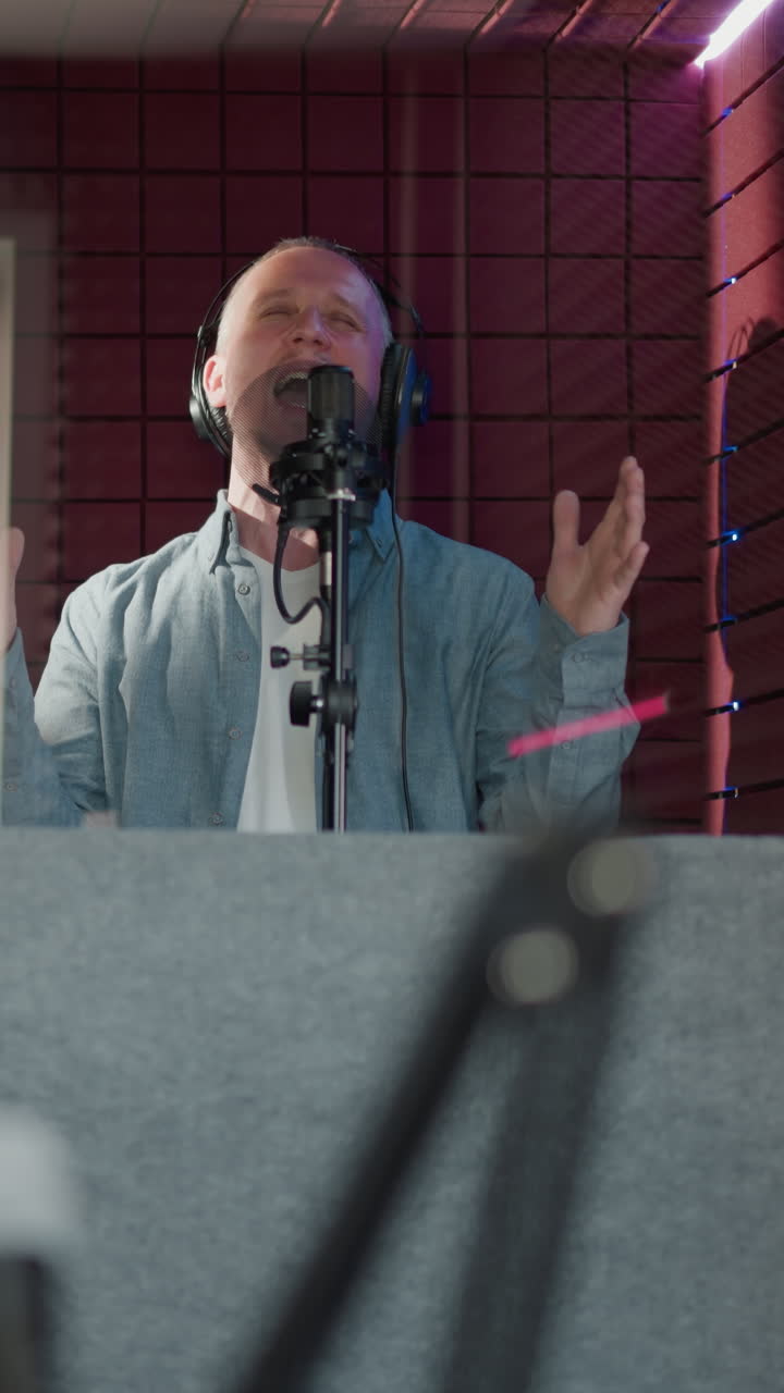 el hombre canta apasionadamente en un estudio de grabación, con auriculares negros y una camisa azul de manga larga. la escena se filma a través de un espejo, un gesto de la mano, en un fondo rojo con paneles acústicos