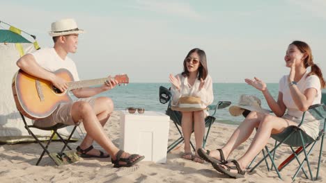 행복한 친구는 캠핑 텐트 근처의 모래 해변에서 휴가를 함께 미소 짓고 캠핑에서 기타를 연주하고 박수를 치며 즐거움을 얻습니다.