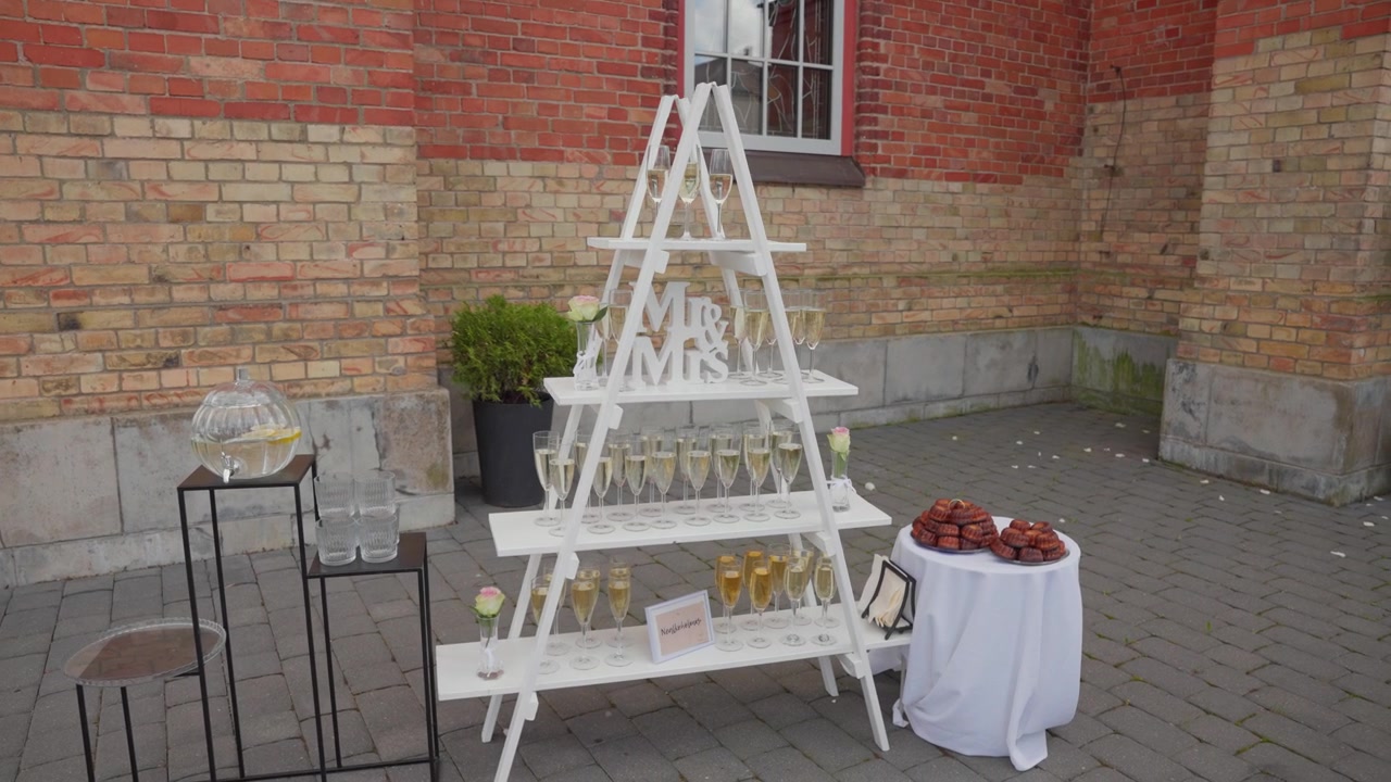 Tasteful wedding drink and dessert station features champagne glasses, donuts, and stylish decor on white stands in a courtyard with rustic brick walls, soft daylight, and inviting setup.