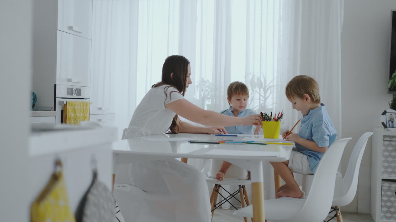 Mom helps sons learn to draw doing homework preschool preparation at home sitting in the white kitchen.