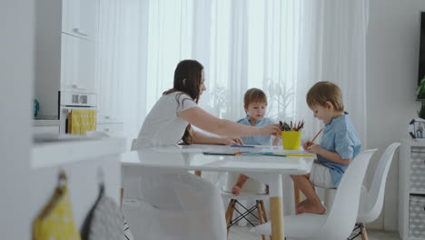 mamá ayuda a los hijos a aprender a dibujar haciendo la tarea de preparación preescolar en casa sentado en la cocina blanca.