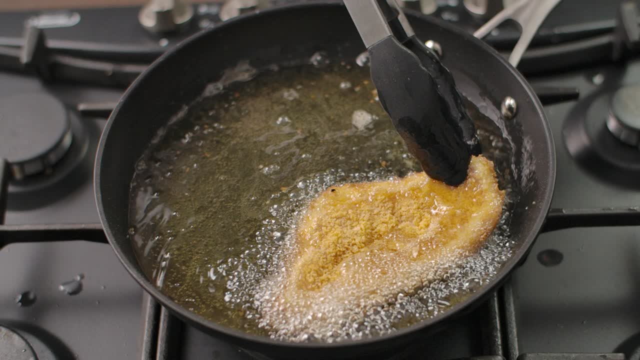Nærbillede i høj vinkel af en perfekt paneret kyllingeschnitzel, der syder i varm olie, løftet med en tang for at afsløre dens sprøde, gyldne tekstur
