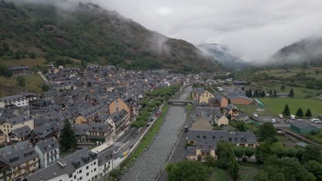 a breathtaking drone shot captures the view of a charming pyrenees mountain town on the spanish side, perched above a tranquil riverbed, enveloped in cozy charm on a cold, cloudy day